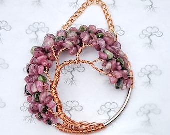 May include: A handcrafted copper wire tree of life wall hanging. The tree features a circular frame and branches adorned with pink and green gemstones. A gold-colored chain is attached for hanging.