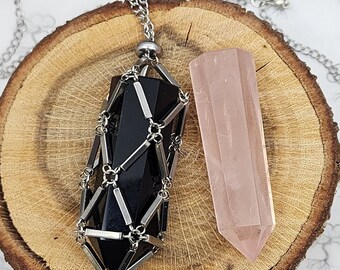 May include: A black crystal encased in a silver cage pendant and a pink crystal point pendant on a silver chain. Both pendants are displayed on a wooden slice.