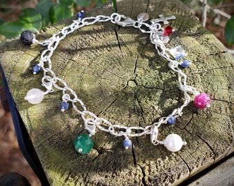 May include: A silver charm bracelet with a variety of charms, including a pink stone, a green stone, a blue stone, a pearl, a lava rock, and a cross. The bracelet is on a wooden surface.