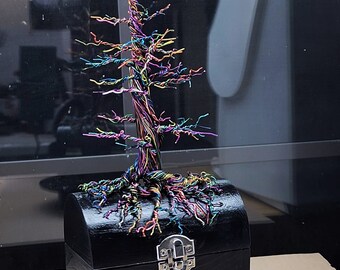 May include: A handcrafted wire tree sculpture with a rainbow finish, mounted on a small, black wooden chest. The tree's branches and trunk are intricately twisted, showcasing a spectrum of colors. The chest has a silver clasp.