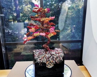 May include: A handcrafted wire tree sculpture with a colorful canopy of red, pink, yellow, and orange. The tree sits in a black wooden box adorned with clear crystals and a silver clasp. The sculpture is displayed on a mirrored surface.