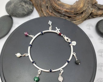 May include: A silver charm bracelet with a variety of charms, including a cross, a pearl, a red stone, a green stone, a pink stone, and a black lava rock. The bracelet is decorated with small blue beads.