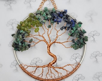 May include: A handcrafted tree of life wall hanging. The tree features a copper wire design with a silver metal ring and a gold chain for hanging. The tree's leaves are made of green, blue, and dark green gemstones.