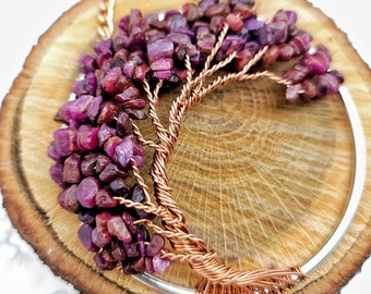 May include: A handcrafted necklace featuring a copper wire tree design, adorned with vibrant, deep red ruby chips. The tree is set against a silver-toned crescent and a wooden background, creating a natural and artistic aesthetic.