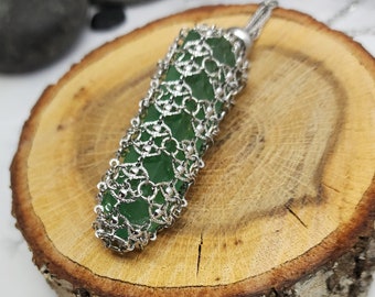May include: A green gemstone pendant encased in a silver wire mesh, resting on a wooden surface.