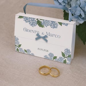 Può includere: Una scatola regalo bianca con manico a nastro blu, decorata con motivi floreali blu e i nomi "Ginevra & Marco" e la data "15.05.2026". Due fedi nuziali d'oro in primo piano, con fiori blu sullo sfondo.