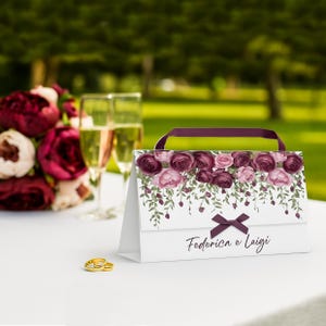 Puede incluir: Una bolsa de regalo blanca con un diseño floral con rosas burdeos y rosas y hojas verdes. Tiene un asa y un lazo burdeos. Los nombres "Federica e Luigi" están impresos en la parte delantera. Dos anillos de boda dorados están en primer plano.
