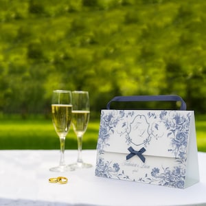 Puede incluir: Una bolsa de regalo blanca con estampados florales azules, un lazo azul y un asa azul está sobre una superficie blanca. Dos copas de champán y dos anillos de boda dorados también están sobre la mesa. El fondo es verde borroso.