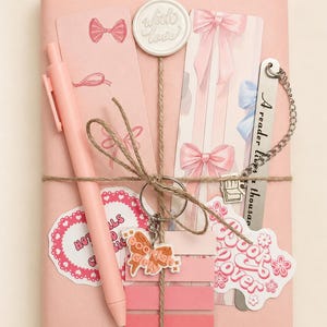 Könnte beinhalten: Ein rechteckiges Paket in rosa Papier, verschnürt mit Bindfaden und einem Wachssiegel mit der Aufschrift With Love. Es enthält einen rosa Stift, zwei Lesezeichen mit Schleifenmotiven, ein Metall-Lesezeichen mit Kette, einen Buch-Anhänger und Aufkleber.