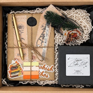 May include: A gift box containing a brown paper-wrapped book with a gold pen, a black wax seal, and two bookmarks. One bookmark reads "JUST ONE MORE CHAPTER." Includes a black box labelled "Lemon Cake Roll," stickers, and a pine sprig.