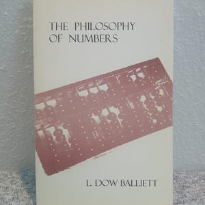 May include: A softcover book titled The Philosophy of Numbers by L. Dow Balliett. The off-white cover features a dark red graphic illustration of an abacus positioned diagonally across the centre, with black text printed above and below it.