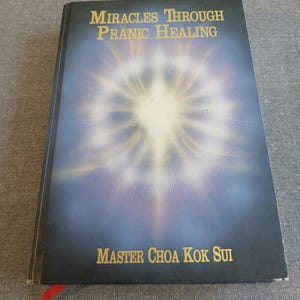May include: A hardcover book titled "Miracles Through Pranic Healing" by Master Choa Kok Sui. The cover features a blue background with a bright, radiating light design. A red bookmark is visible.