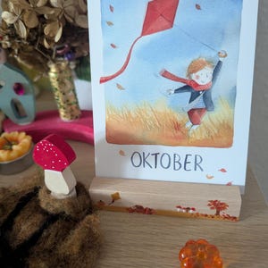 Könnte beinhalten: Aquarellillustration eines Kindes, das einen roten Drachen auf einem Feld steigen lässt, mit dem Wort "OKTOBER" am unteren Rand. Das Bild wird auf einem Holzständer angezeigt. Weitere dekorative Elemente sind ein Holzpilz und ein kleiner orangefarbener Glasgegenstand.