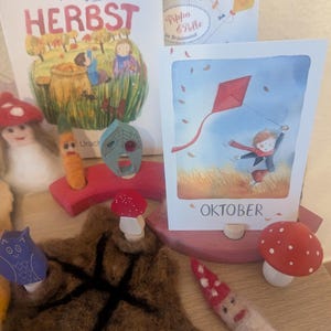 以下が含まれることがあります： 子供向けの絵本と木製おもちゃのコレクション。「HERBST」という本が見え、赤い凧を揚げている子供の写真もあります。キノコとフクロウの置物、フェルトの葉もあります。