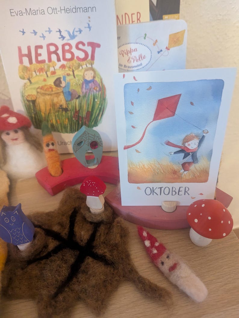 以下が含まれることがあります： 子供向けの絵本と木製おもちゃのコレクション。「HERBST」という本が見え、赤い凧を揚げている子供の写真もあります。キノコとフクロウの置物、フェルトの葉もあります。