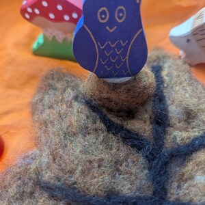 以下が含まれることがあります： 金色のアクセントが付いたネイビーブルーの木製のフクロウの置物が、フェルトの木の幹の上にあります。白い水玉模様の赤いキノコと緑色のベースが背景にあります。遊び心のある森のテーマです。