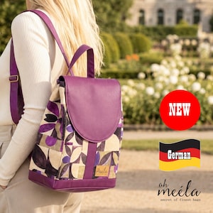 Könnte beinhalten: Ein violetter und beiger Rucksack mit geometrischem Blumenmuster, einer einfarbigen violetten Klappe und violetten Schultergurten. Ein kleines rechteckiges Etikett mit dem Text oh meila ist vorne angebracht. Der Rucksack wird auf dem Rücken getragen.
