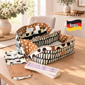 May include: A rectangular fabric organiser with a black and cream abstract pattern and brown polka dot interior lining. It features multiple internal compartments, a zippered centre section, and a detachable flap with elastic loops and a mesh pocket.