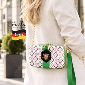 May include: A rectangular crossbody bag featuring a white base with a repeating maroon clover pattern, a central green vertical stripe, and a black hexagonal patch with a gold bee emblem. It includes a gold chain strap and a green wrist strap.