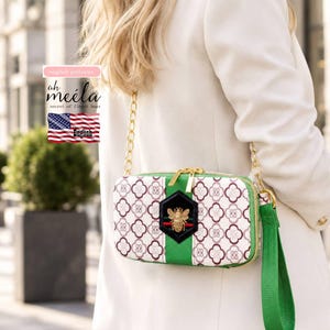 May include: A rectangular crossbody bag featuring a white and maroon quatrefoil pattern, a central green vertical stripe, and a black hexagonal patch with a gold bee emblem. The bag includes a gold chain strap and a green wristlet strap.
