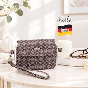 May include: A rectangular wristlet bag featuring a dark brown fabric with a repeating light-colored floral pattern, a front flap with a small silver-toned bow accent, a white zipper trim, and a matching wrist strap. The text oh meila secret of finest bags is visible.