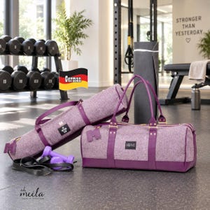 Könnte beinhalten: Eine Yogamatten-Tasche aus violettem meliertem Stoff und eine passende Sporttasche mit dunkelvioletten Besätzen und goldfarbenen Beschlägen. Die Sporttasche hat ein schwarzes rechteckiges Etikett mit weißem Text und die Yogatasche trägt ein M.