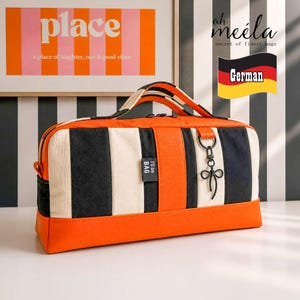 Könnte beinhalten: Eine rechteckige Tasche mit vertikalen Streifen in Orange, Schwarz und Cremeweiß. Die Tasche hat einen orangefarbenen Besatz an der Basis und an den Griffen, ein schwarzes Stoffetikett mit dem Text "I'll be BAG" und einen schwarzen Metallanhänger in Schleifenform.