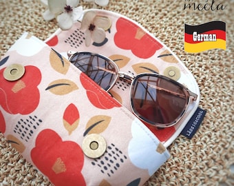 Puede incluir: Un estuche de tela para gafas con fondo rosa claro y un estampado de flores rojas y blancas, hojas marrones y rayas negras. El estuche cuenta con un cierre magnético dorado y una etiqueta lateral negra con el texto LÄSSIG&COOL.