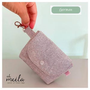 Könnte beinhalten: Eine kleine, hellrosa Stofftasche mit Klappenverschluss und Druckknopf. Sie hat eine kleine rosa Lasche an der Seite und einen silberfarbenen Verschluss. Der Text "oh meela secret of finest bags" ist unten links aufgedruckt.
