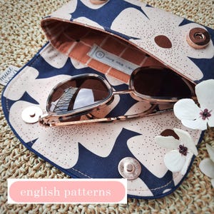Könnte beinhalten: Sonnenbrille in einem geöffneten Etui mit blau-weißem Blumenmuster. Das Etui hat einen Druckknopfverschluss und ein Innenfutter in Pfirsichfarbe. Die Sonnenbrille hat ein Schildpattgestell und braune Gläser. Der Text "english patterns" ist sichtbar.
