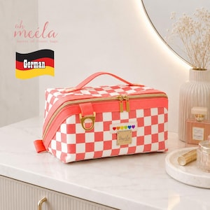 Può includere: Una trousse rettangolare con motivo a scacchi color corallo e bianco. Presenta un manico superiore corallo, cerniere color oro e una targhetta con logo in metallo dorato. Una piccola toppa bianca con cuori colorati è centrata sul davanti.
