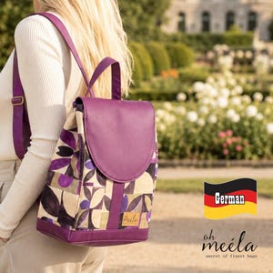 Könnte beinhalten: Ein violetter und beiger Rucksack mit geometrischem Blumenmuster, einer einfarbigen violetten Klappe und passenden violetten Schultergurten. Ein kleines rechteckiges Etikett mit dem Text oh meila ist vorne am Rucksack nahe der Basis angebracht.