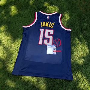 Może przedstawiać: Granatowy koszykarski trykot z napisem "JOKIC" w złotych literach i numerem "15" w kolorze białym z czerwonym obramowaniem. Trykot ma białe i bordowe wykończenie wokół szyi i otworów na ramiona. Dołączona jest przezroczysta plastikowa osłona.