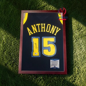 May include: Framed black basketball jersey with the name "ANTHONY" and the number "15" in yellow. The jersey has a yellow trim and a signature. The frame is dark red.