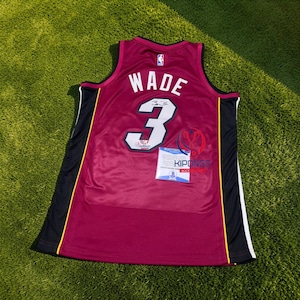 Może przedstawiać: Bordowa koszulka do koszykówki z napisem "WADE" i numerem "3" w kolorze białym. Koszulka ma czarne i żółte akcenty po bokach oraz małe logo NBA. Dołączony jest certyfikat autentyczności.