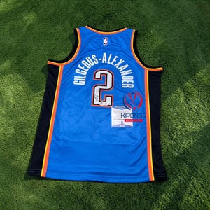 Może przedstawiać: Niebieska koszulka do koszykówki z napisem "GILGEOUS-ALEXANDER" i numerem "2" w kolorze białym. Koszulka ma czarne i pomarańczowe akcenty oraz logo NBA. Do koszulki dołączona jest mała metka z tekstem.