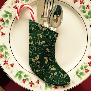 May include: A festive Christmas table setting featuring a white plate with a holly and berry design, a red charger, and silverware. A small green stocking with gold holly accents holds the silverware and a candy cane.