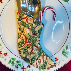 May include: A festive Christmas table setting featuring a white plate with a holly and berry design. Silverware, including a knife, fork, and spoon, is nestled in a decorative fabric stocking. A candy cane adds a touch of holiday cheer.