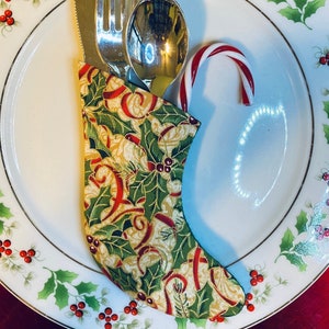 May include: A festive Christmas table setting featuring a white plate with a holly and berry design. Silverware is nestled in a fabric stocking with a holly and ribbon pattern. A candy cane adds a touch of holiday cheer.