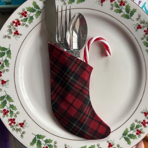 May include: A festive Christmas table setting featuring a white plate with a holly and berry design. Silverware is nestled in a red and black plaid stocking, with a red and white candy cane on the plate. The plate has a gold rim.