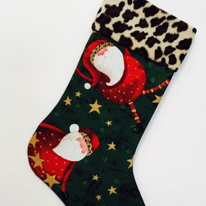 May include: Christmas stocking with a green background featuring Santa Claus illustrations and gold stars. The cuff has a leopard print design. A red ribbon is attached for hanging.