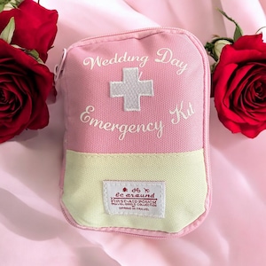 Può includere: Un kit di pronto soccorso in tela rosa con il testo "Wedding Day Emergency Kit" ricamato sul davanti. Il kit ha una cerniera gialla e una croce bianca ricamata sul davanti. Il kit è su una superficie rosa.