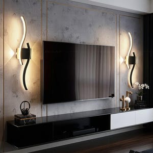May include: Contemporary wall lights with a black finish and a curved, illuminated design. The lights are mounted on a textured grey wall, either side of a large flat-screen television. The room features a black and white entertainment unit.