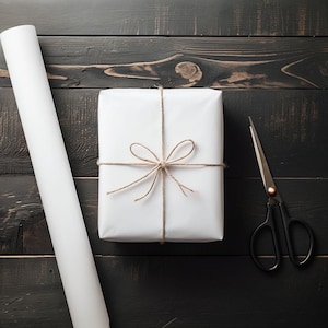 May include: A white gift box wrapped with twine and a bow. The box is sitting on a dark wood surface with a roll of white wrapping paper and a pair of scissors.