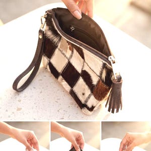 May include: A rectangular wristlet pouch featuring a patchwork design of brown, black, and white cowhide fur. The accessory includes a dark brown leather wrist strap, a matching leather tassel zipper pull, and a dark brown interior lining.