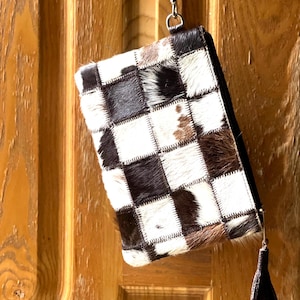 Puede incluir: Bolso de mano rectangular con diseño de patchwork. La bolsa presenta cuadrados de piel de animal negra, blanca y marrón. Una borla negra cuelga de la cremallera y una correa de cuero está sujeta a la parte superior. La bolsa está colgada de una puerta de madera.