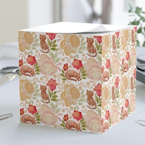 May include: A white notepad cube with a repeating watercolour pattern. The design features stylized faces, cats, flowers, and foliage in shades of peach, red, and green. The notepad is on a white surface.