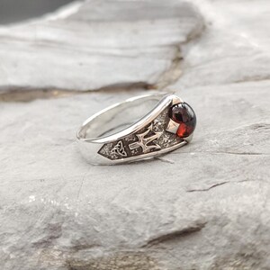 May include: A silver ring featuring a polished dark red cabochon gemstone set into a textured band. The metal surface displays an engraved trident symbol alongside a small triquetra knot design, creating a detailed metallic finish on the band.