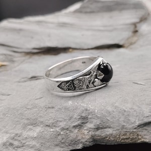 May include: A silver ring with a black gemstone and wolf design. The ring features a round black stone and detailed wolf and Celtic knot engravings on the band. The ring is displayed on a gray stone surface.