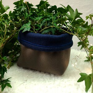 Peut inclure: Une plante verte en pot avec du lierre qui pousse dans un pot en tissu brun et bleu. Le pot a une bordure en tissu bleu et repose sur une surface blanche et moelleuse.